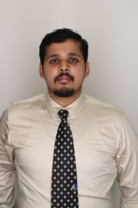A man with short dark hair, a beard, and mustache is wearing a cream-colored shirt and a patterned tie, standing against a plain light background.