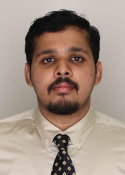 A man with short dark hair, a beard, and mustache is wearing a cream-colored shirt and a patterned tie, standing against a plain light background.