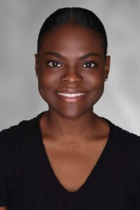 A person with short hair is smiling at the camera, wearing a black top, against a neutral gray background.