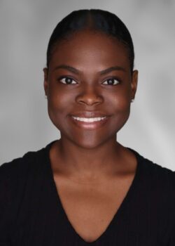 A person with short hair is smiling at the camera, wearing a black top, against a neutral gray background.