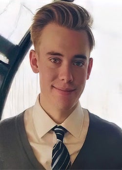 A young man with light brown hair wearing a white shirt, striped tie, and gray sweater smiles at the camera in a well-lit indoor setting.