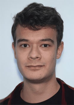 A young man with short brown hair, light skin, and a neutral expression looks directly at the camera against a plain light background.