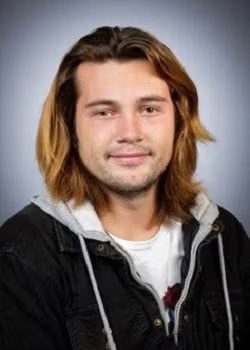 A young man with shoulder-length brown hair, wearing a black jacket over a gray hoodie and white t-shirt, poses against a plain, gradient background.
