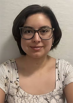 A person with short dark hair and glasses is wearing a light-colored patterned top and is facing the camera, seated against a plain light background.