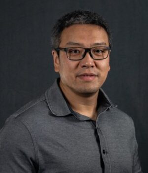 A man with short dark hair and glasses, wearing a grey collared shirt, poses in front of a plain dark background.