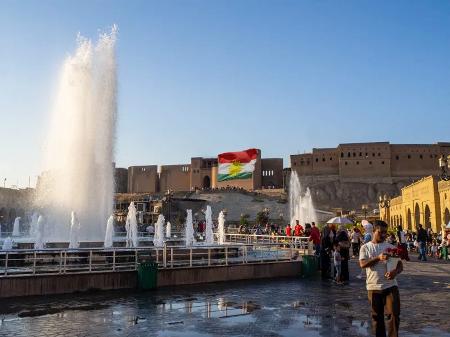 Fountains in a public square with people walking. A large flag is displayed on a building in the background under a blue sky.