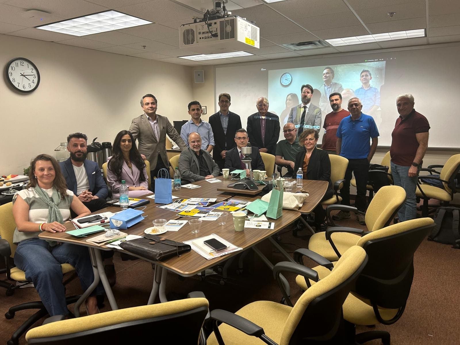 A group of people pose for a photo in a conference room around a table with laptops, notebooks, and refreshments; a video call is projected on the wall behind them.