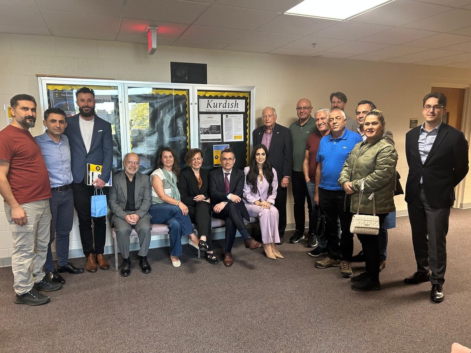 A group of people poses together indoors near a bulletin board labeled "Kurdish," with some seated on a bench and others standing behind them.