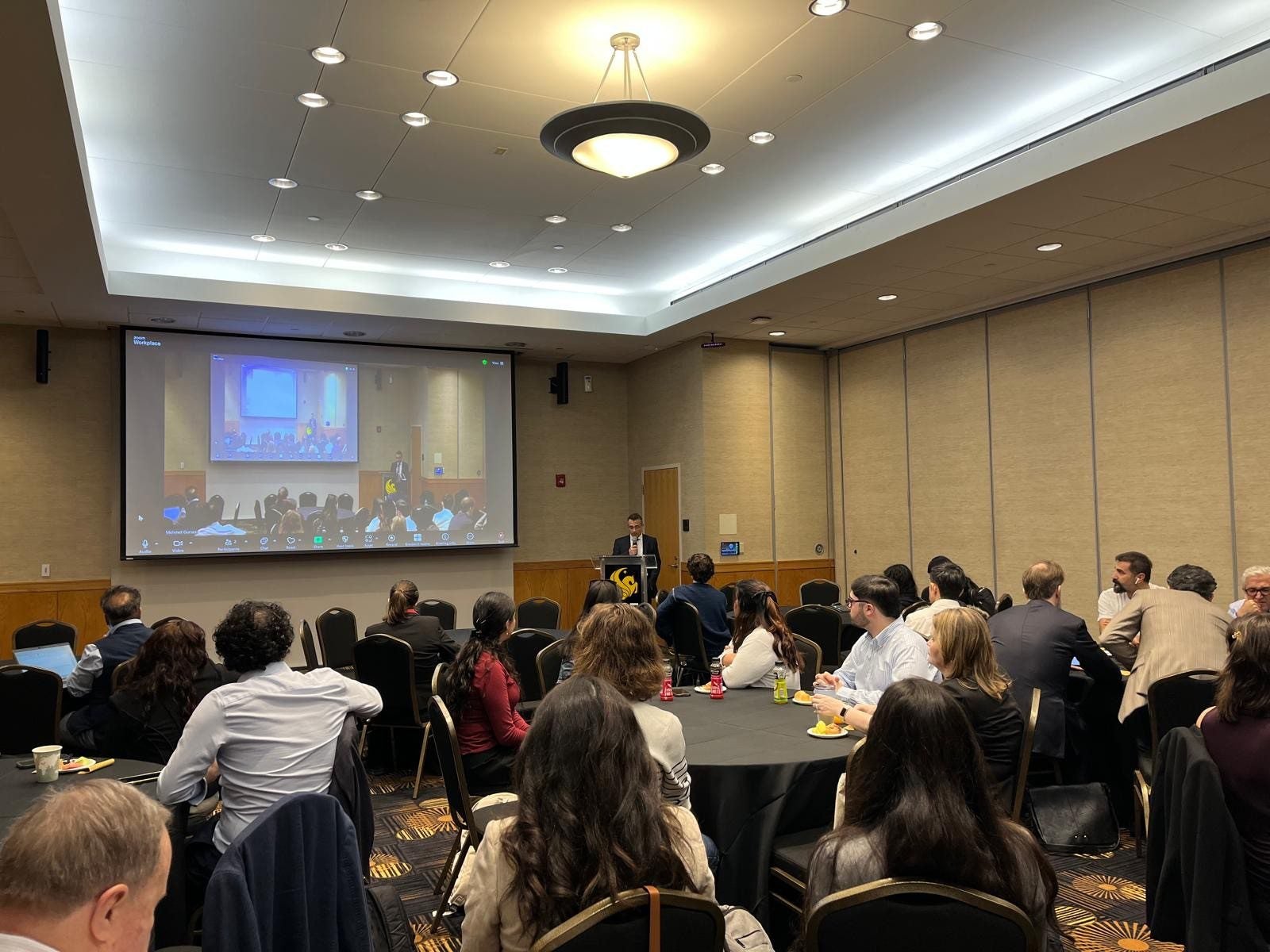 People seated at round tables in a conference room watch a speaker at a podium with a large screen displaying a presentation at the front of the room.