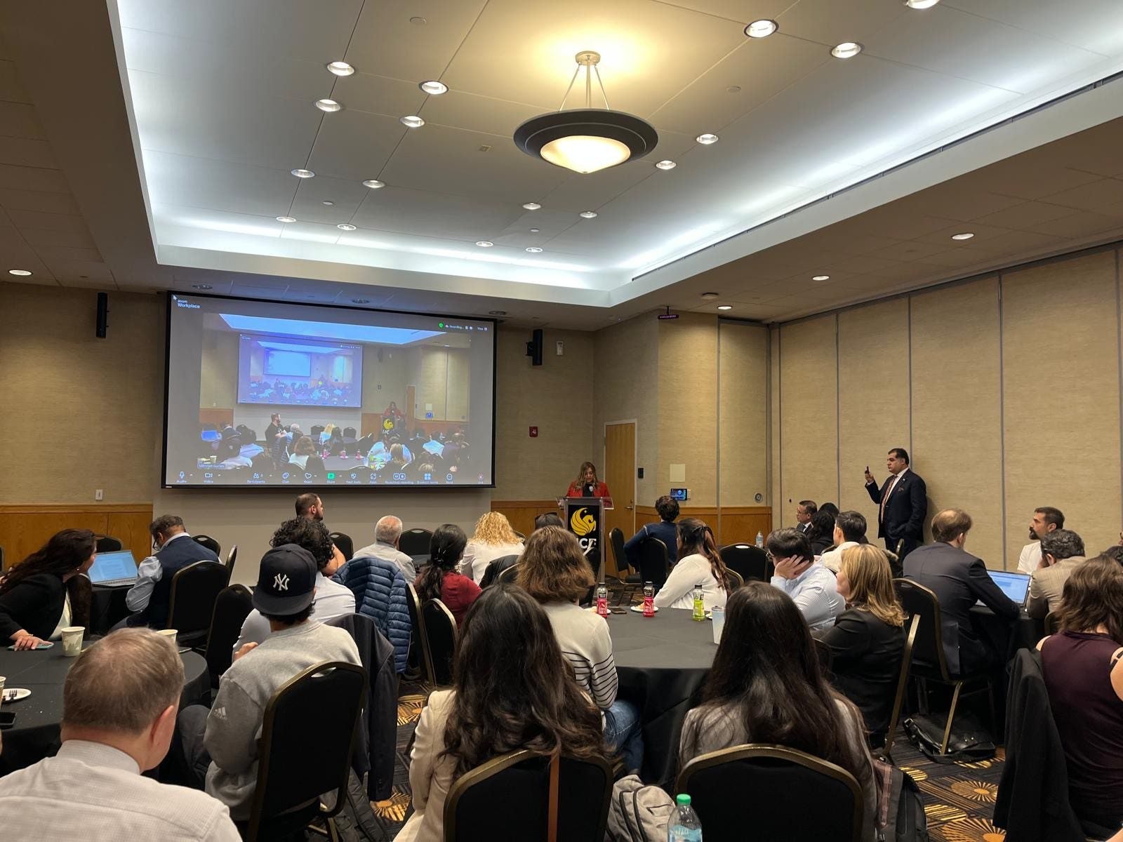 A conference room with people seated at round tables, listening to a speaker at a podium; a large screen displays a live video of the event.