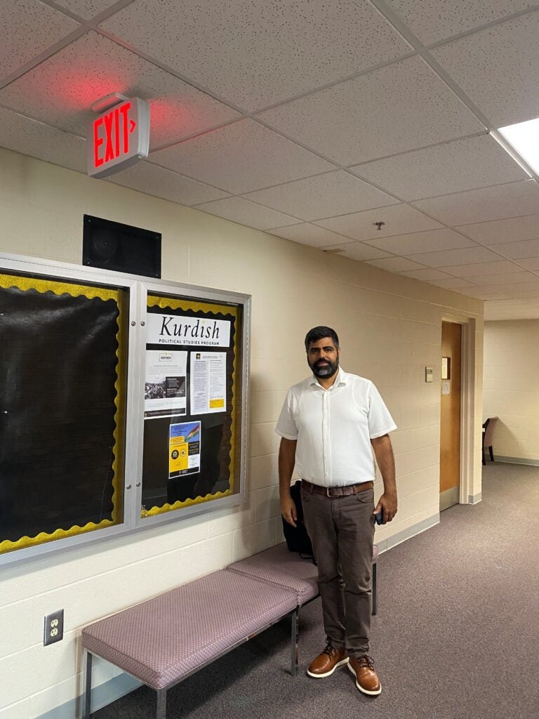 A man in a white shirt stands next to a bulletin board labeled "Kurdish" in a hallway with an EXIT sign above.
