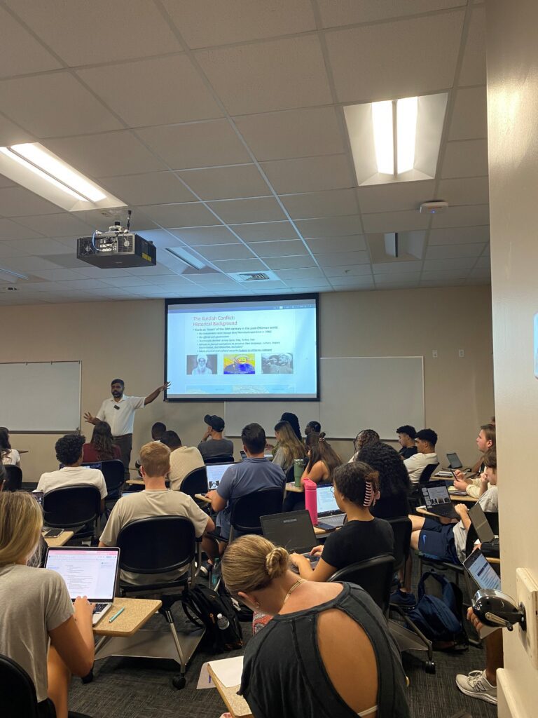 A classroom of students sits at desks with laptops open while a teacher stands at the front pointing to a presentation slide projected on a screen.