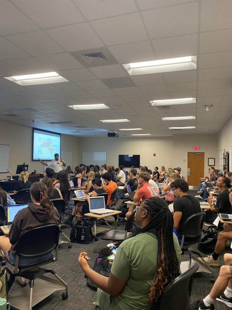 A classroom filled with students seated at desks, some using laptops, listening to an instructor presenting slides at the front of the room.
