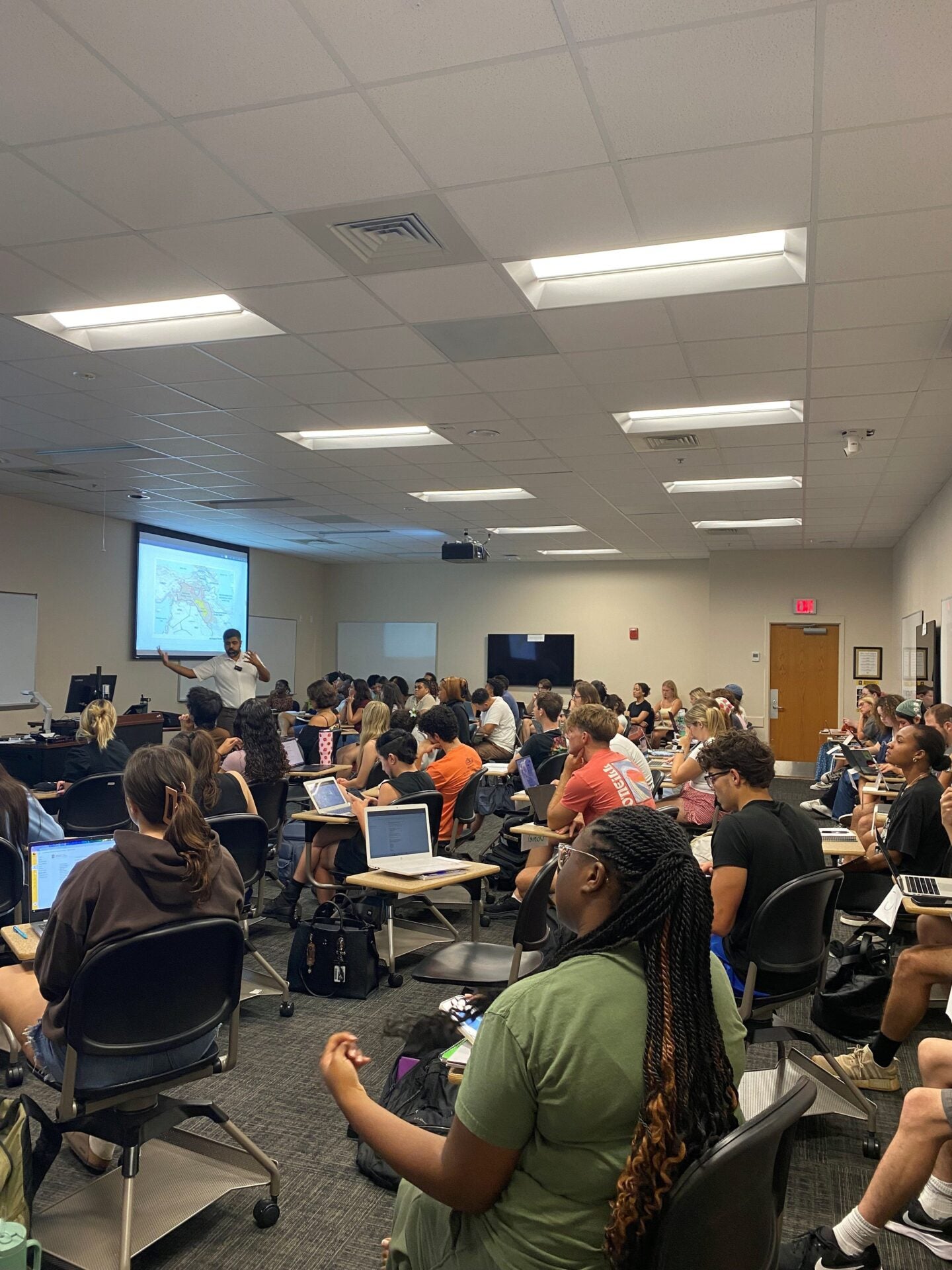 A classroom filled with students sitting at desks, some using laptops, while an instructor at the front presents a lesson with a projected image on the screen.