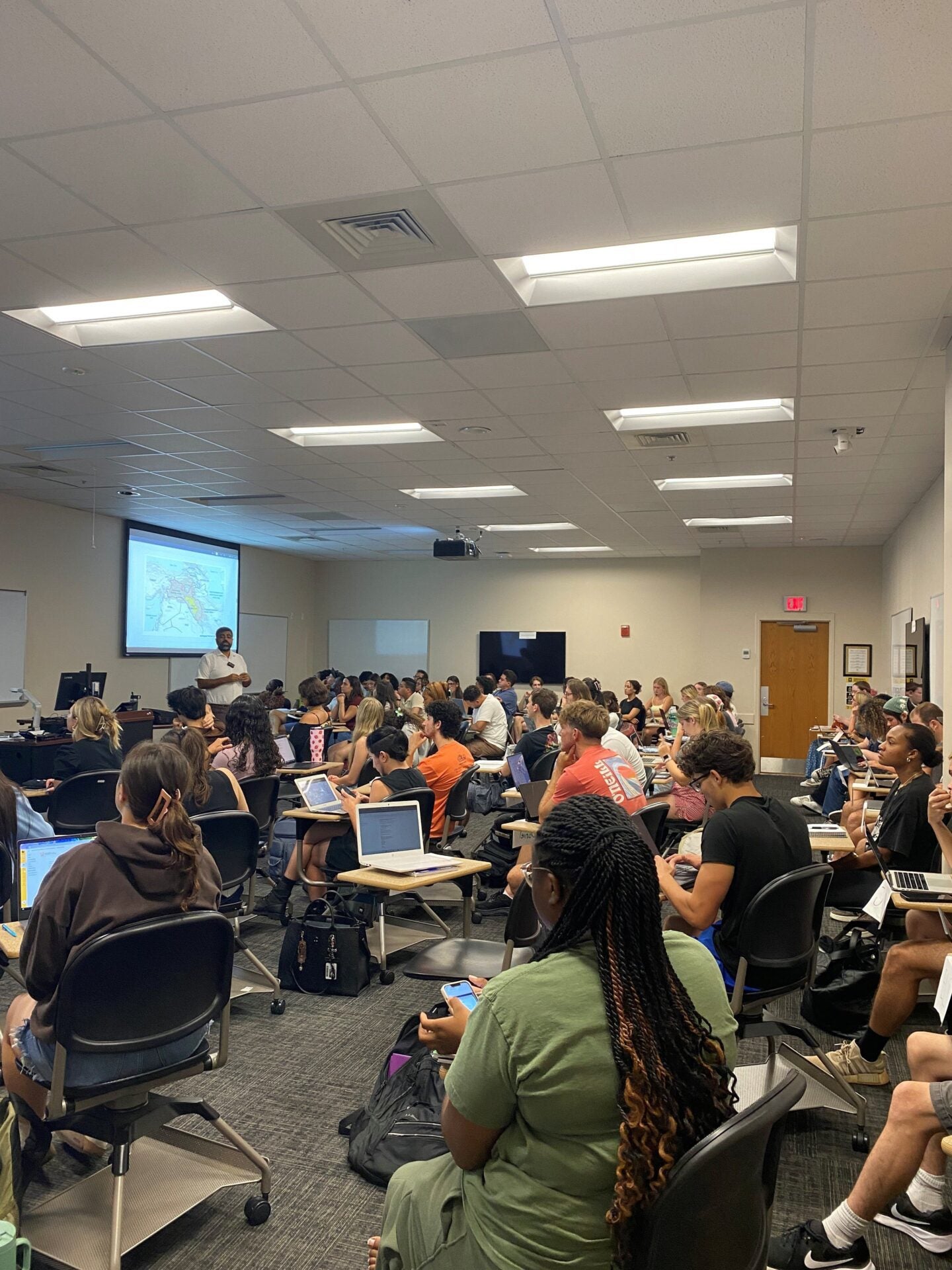 A classroom full of students sits at desks with laptops open, facing a professor who is giving a presentation in front of a projected screen.