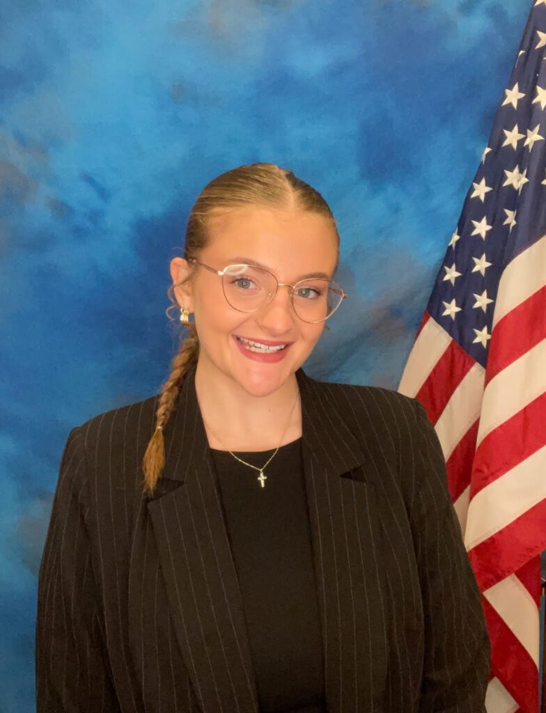 A young woman in a black pinstripe suit and glasses stands in front of a blue backdrop next to an American flag, smiling at the camera.