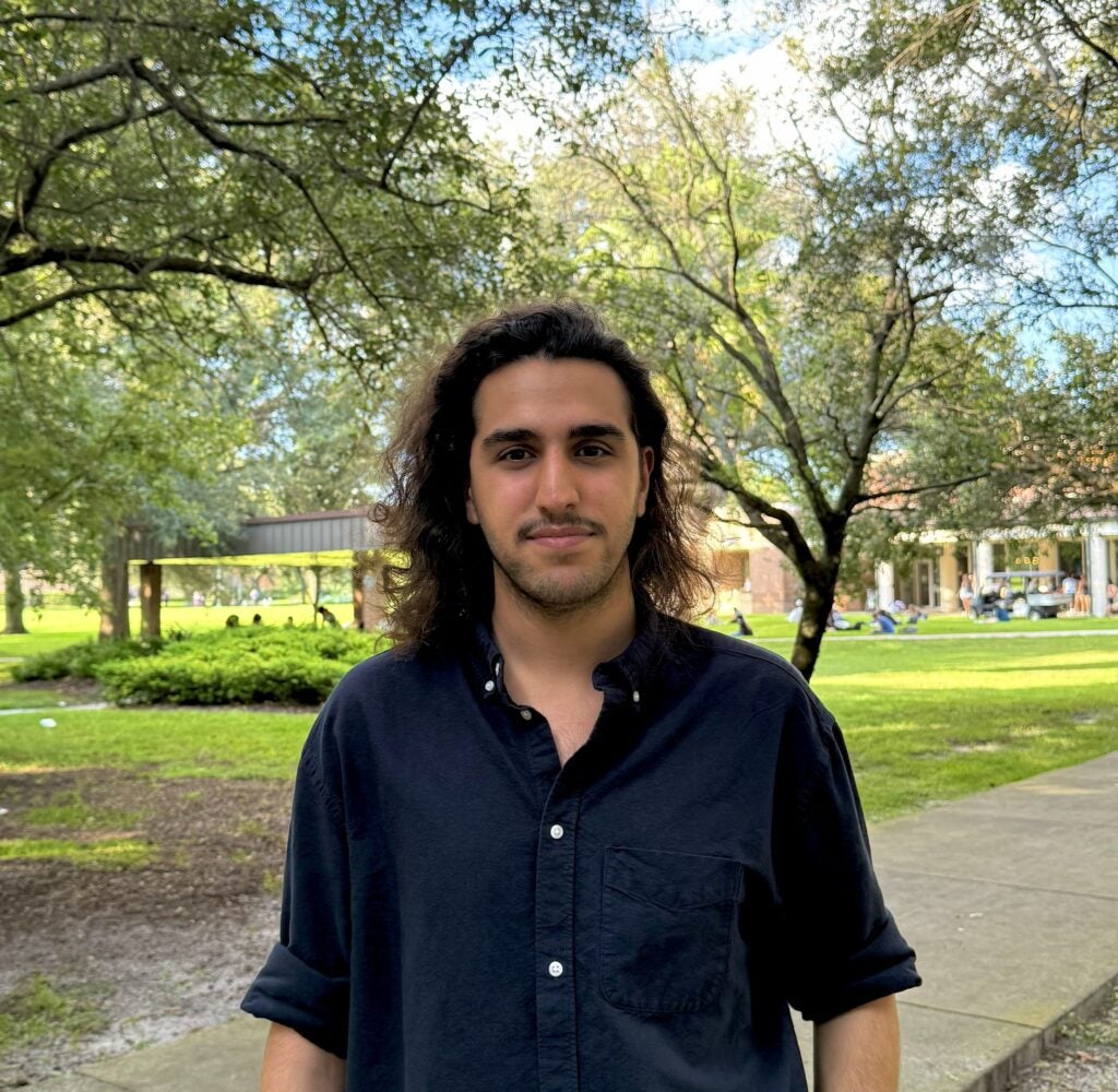 A young man with long, dark, wavy hair and a mustache stands in front of a green lawn with trees and buildings in the background.