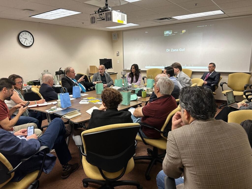 A group of people are seated around a conference table, listening to a presentation displayed on a projection screen.