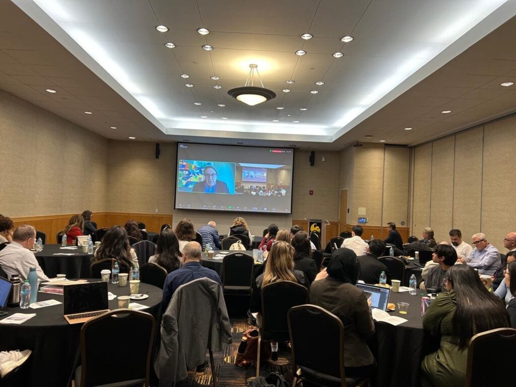 A presenter appears on a large screen in a conference room where attendees sit at round tables.