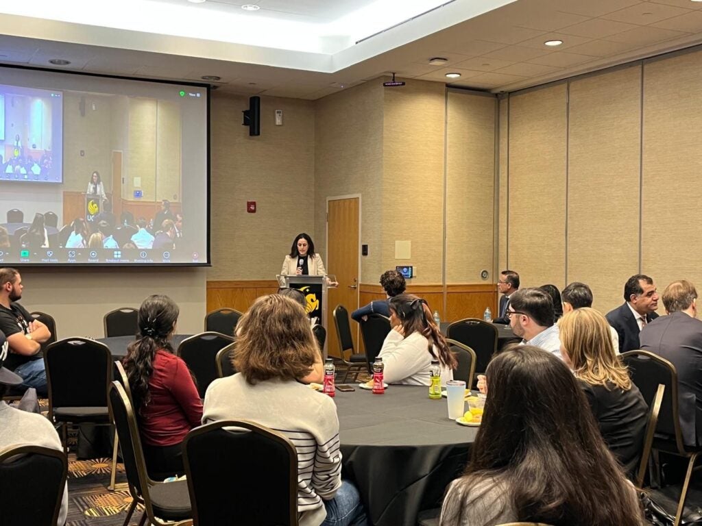 A woman speaks at a podium in front of a large screen displaying a virtual presentation. An audience sits at round tables facing her.