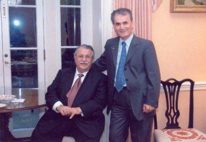 Two men in suits pose indoors; one is seated at a round table, the other stands beside him. The setting appears formal with light-colored walls and a window in the background.