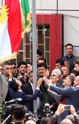A group of people raise a flag outdoors in front of a crowd, with cameras in the foreground capturing the event.