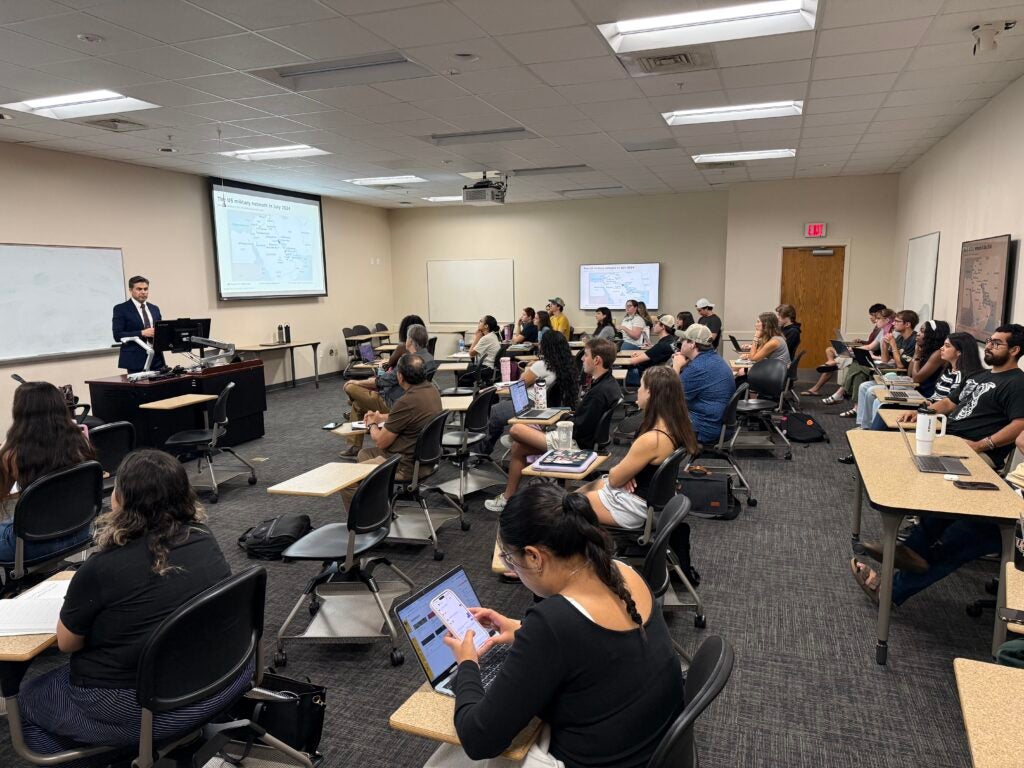A professor lectures to a classroom of students seated at desks, with a map of US military bases projected on the screen.