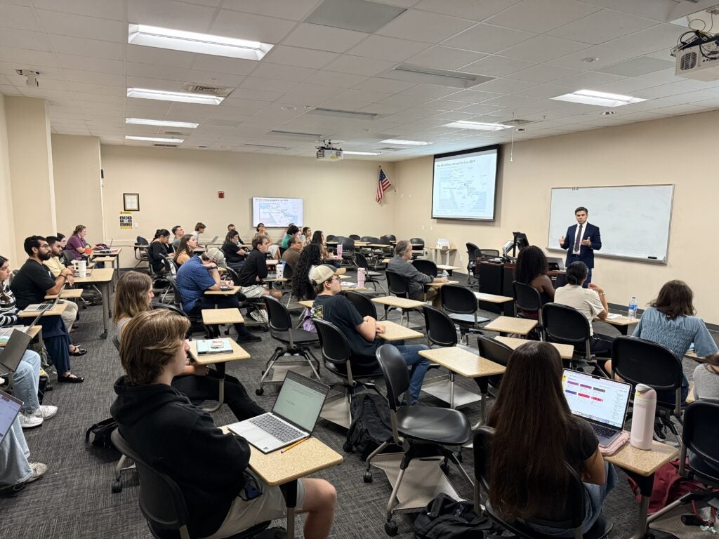 A lecturer in a suit stands at the front of a classroom, presenting to a group of students seated at desks with laptops.