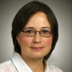 A woman with short dark hair and glasses is wearing a white patterned blouse and a blue scarf. She is facing the camera against a plain background.