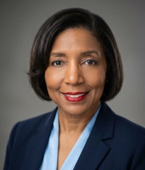 A woman with short dark hair wearing a navy blazer and light blue blouse, smiling at the camera against a plain gray background.