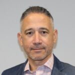 A man with short gray hair and facial stubble is wearing a plaid shirt and a blue blazer, standing against a plain white background.