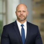 A bald man with a beard, wearing a navy blue suit, white shirt, and blue tie, sits with hands clasped on his lap in a professional setting.