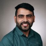A man with short black hair and a beard is smiling, wearing a green button-up shirt, and posing against a plain, neutral background.