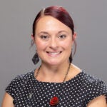 Woman with dark hair pulled back, wearing a black dress with white polka dots and a necklace with a red rose, smiles against a plain gray background.