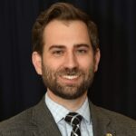 A man with short brown hair and a beard, wearing a gray checked suit, light blue shirt, and a black and white patterned tie, stands in front of a dark background.