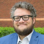 A man with curly hair, a beard, and glasses wearing a blue blazer and striped shirt stands outdoors in front of greenery and a brick wall.