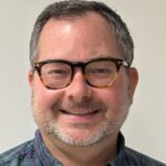 A man with short gray hair, glasses, and a beard smiles at the camera while wearing a green and blue plaid shirt against a plain background.
