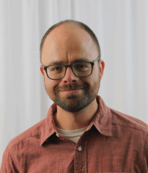 A man with glasses and a beard is wearing a red button-up shirt and standing in front of a white curtain.