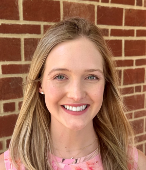 A woman with long blonde hair wearing a pink floral top is smiling in front of a brick wall.
