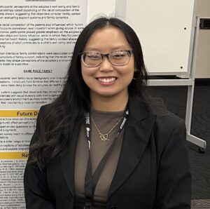 A young woman with glasses, wearing a blazer and a conference lanyard, smiling in front of a research poster.
