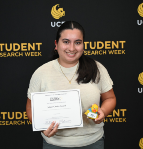 A student stands in front of a UCF Student Research Week backdrop, holding a Judges Choice Award certificate and a small yellow rubber duck.