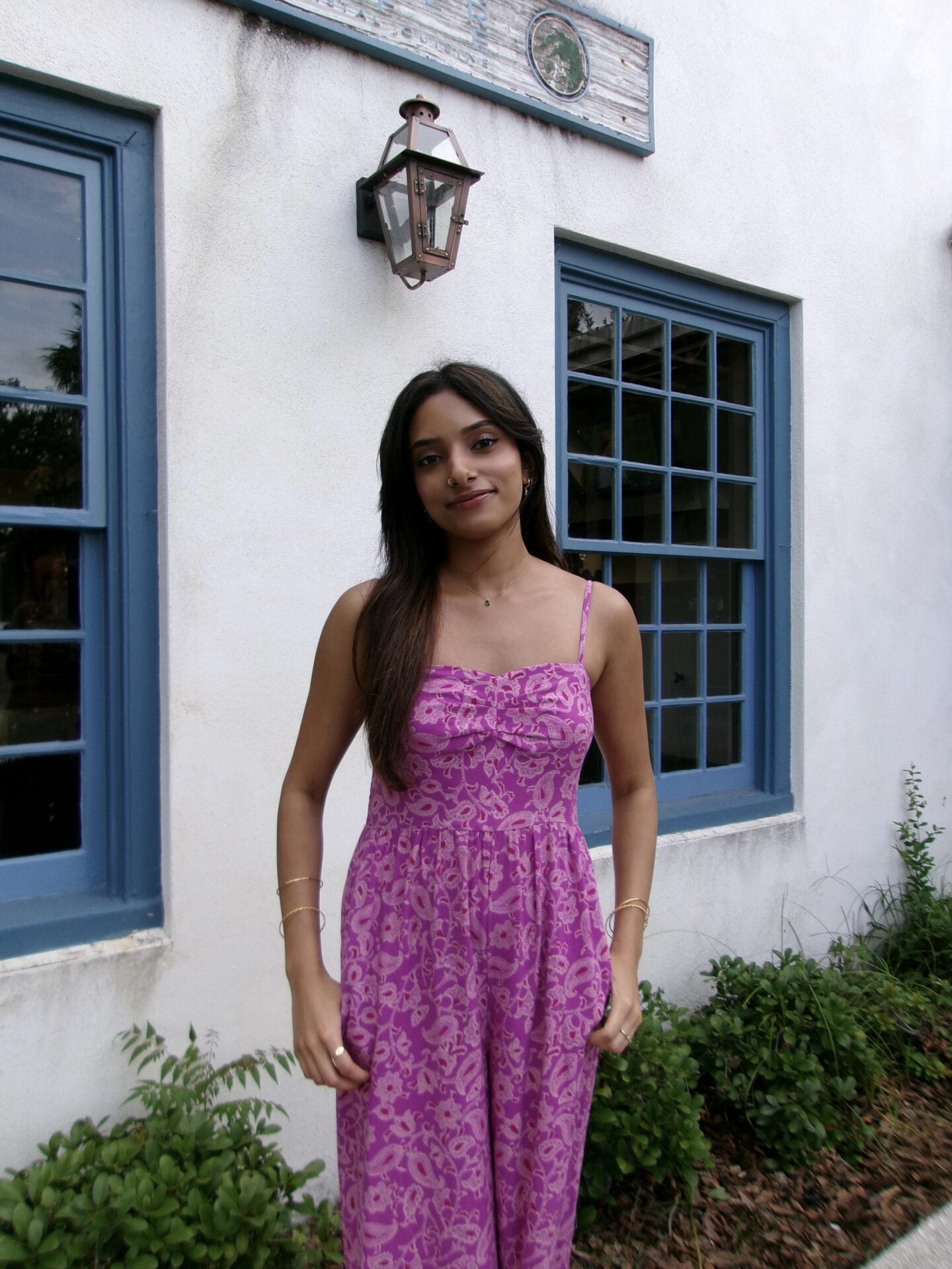 A woman in a purple patterned dress stands in front of a white building with blue-framed windows and a lantern, surrounded by greenery.