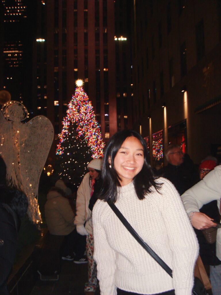 A young woman in a white sweater smiles in front of a decorated Christmas tree at night in a busy outdoor city setting.