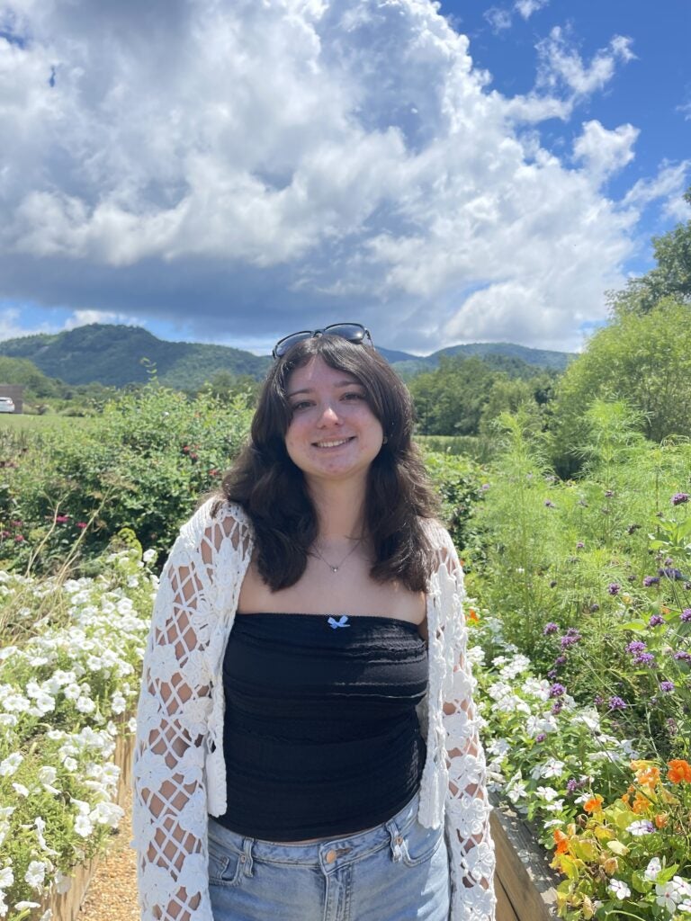 A woman stands in a garden with mountains and a cloudy blue sky in the background. She is wearing a black top, a white crochet cardigan, and sunglasses on her head.