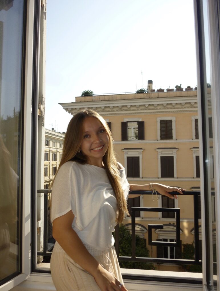 A woman with long hair stands by an open window, smiling, with a beige building and sunny sky visible outside.