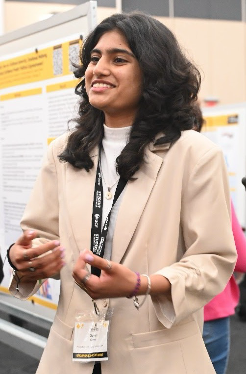 A young woman wearing a beige blazer and conference badge stands in front of a scientific poster, gesturing with her hands while speaking.