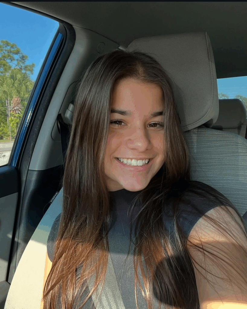 A young woman with long brown hair sits in the passenger seat of a car, smiling at the camera while wearing a seatbelt. Sunlight shines through the window.