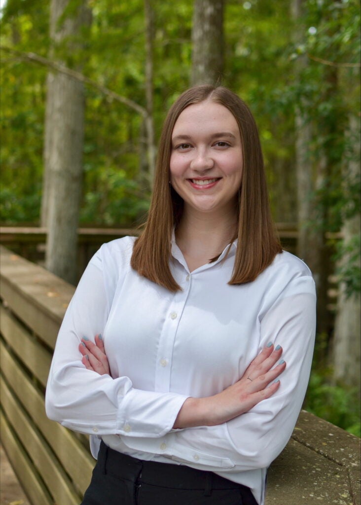 A young woman with straight brown hair, wearing a white button-up shirt, stands outdoors with arms crossed, smiling in front of a wooden railing and trees.