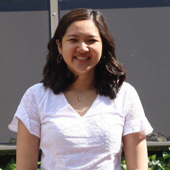 A person with shoulder-length hair is standing outdoors wearing a white eyelet blouse and smiling.