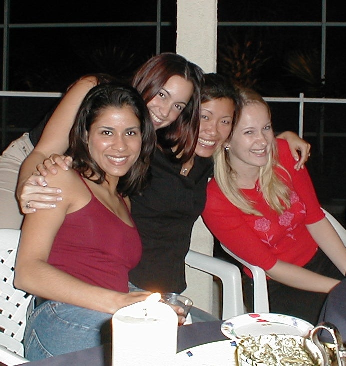 Four women sit closely together, smiling at the camera indoors at a table set with a candle and plates.